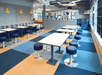 A dining area with two rows of tables with small blue stools at them and another dining and lounge area in the background.