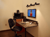 A business center with a desk with a monitor mounted to the wall, and a mini fridge and microwave to the left of it.