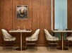 Restaurant interior with two tables and four beige chairs, set with glasses and black napkins, against a wooden wall. A decorative hat is displayed in a frame on the wall.