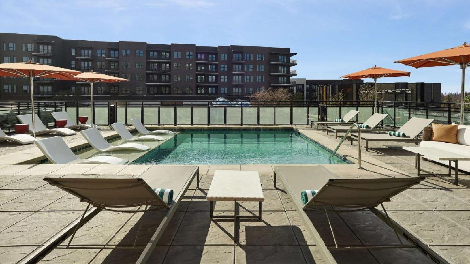 Rooftop pool area with lounge chairs, umbrellas, and a glass railing. Modern apartment buildings in the background.