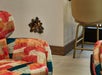 Colorful chairs in a room with tile flooring and cream walls. The wall features a hole shaped like an explosion, revealing what appears to be bricks inside. A barstool is near the window.
