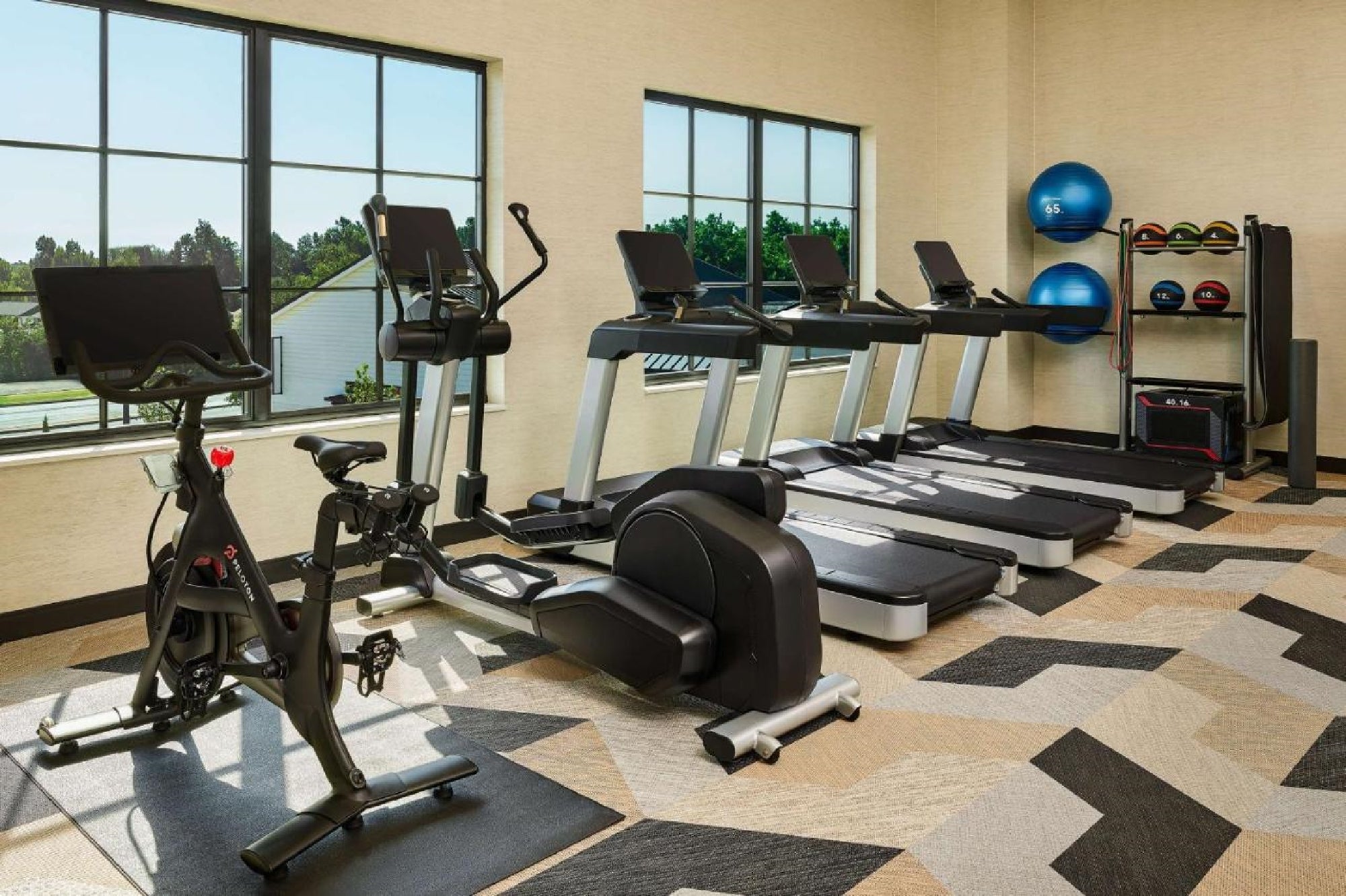 Gym with treadmills, stationary bike, exercise balls, and dumbbells on a patterned floor. Large windows provide natural light.