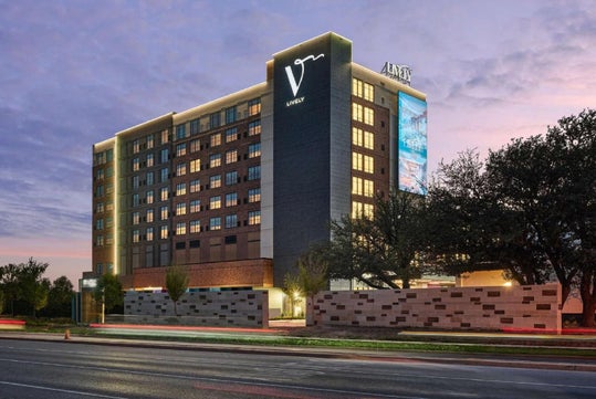 A modern hotel building with illuminated signage stands against a dusk sky. Visible are eight stories, a main road with light trails, trees, and a low wall.