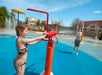 Two children play with water features at an outdoor splash pad. One child operates a red water sprayer while the other plays nearby.