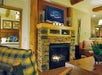 A cozy living room with a stone fireplace, TV mounted above, and wooden beams. A plaid armchair and a wooden coffee table are in the foreground. Bedroom visible through an open door.