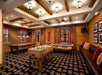 Game room with foosball, shuffleboard, and pool tables. Wood paneling and ceiling beams, patterned carpet, and red accented curtains and cushions create a cozy atmosphere.