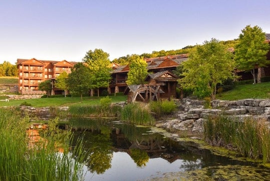 Lodge-style buildings surrounded by trees and a pond with rocks and greenery in the foreground.