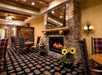 Cozy dining area with wooden beams, stone fireplace, patterned carpet, and plaid chairs. Sunflowers and a large mirror sit above the mantel.