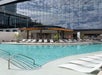 Outdoor hotel pool area with lounge chairs, in-water loungers, and a covered bar, set against a modern building with reflective glass windows.