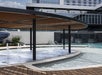 A modern outdoor pool area features a shaded structure, water jets, a curved wooden deck, and a multi-story building in the background.