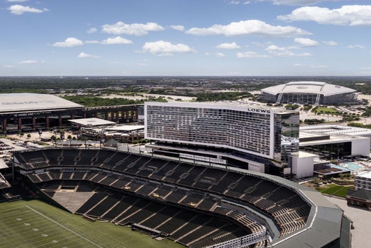 Aerial view of a large outdoor stadium, a nearby hotel, and another domed stadium in the background under a blue sky with scattered clouds.