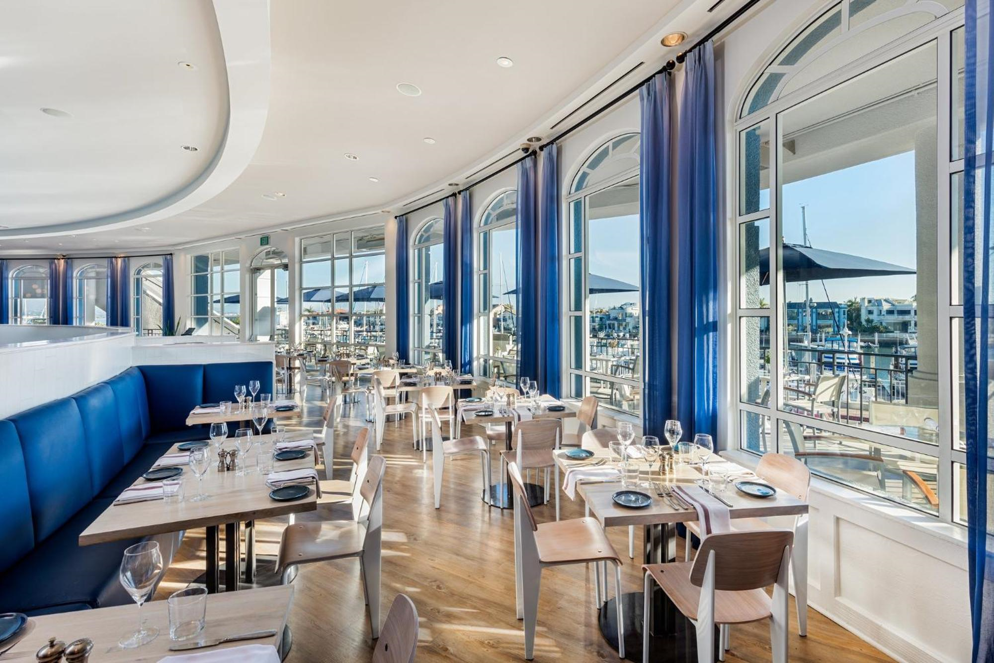 Modern restaurant interior with blue accents, light wooden tables and chairs, large arched windows, and views of an outdoor patio and marina.