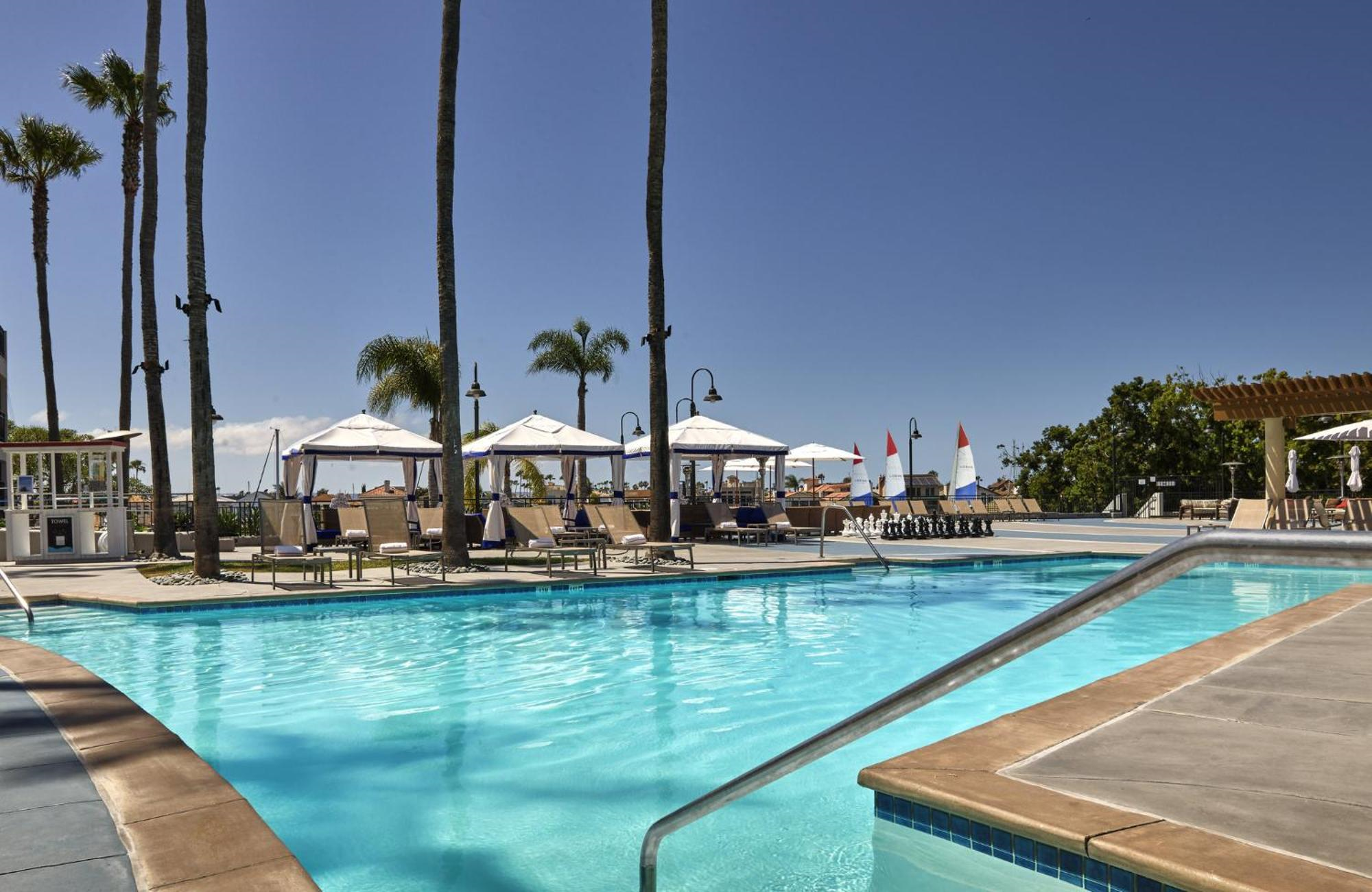 Outdoor swimming pool surrounded by lounge chairs, umbrellas, and palm trees under a clear blue sky.