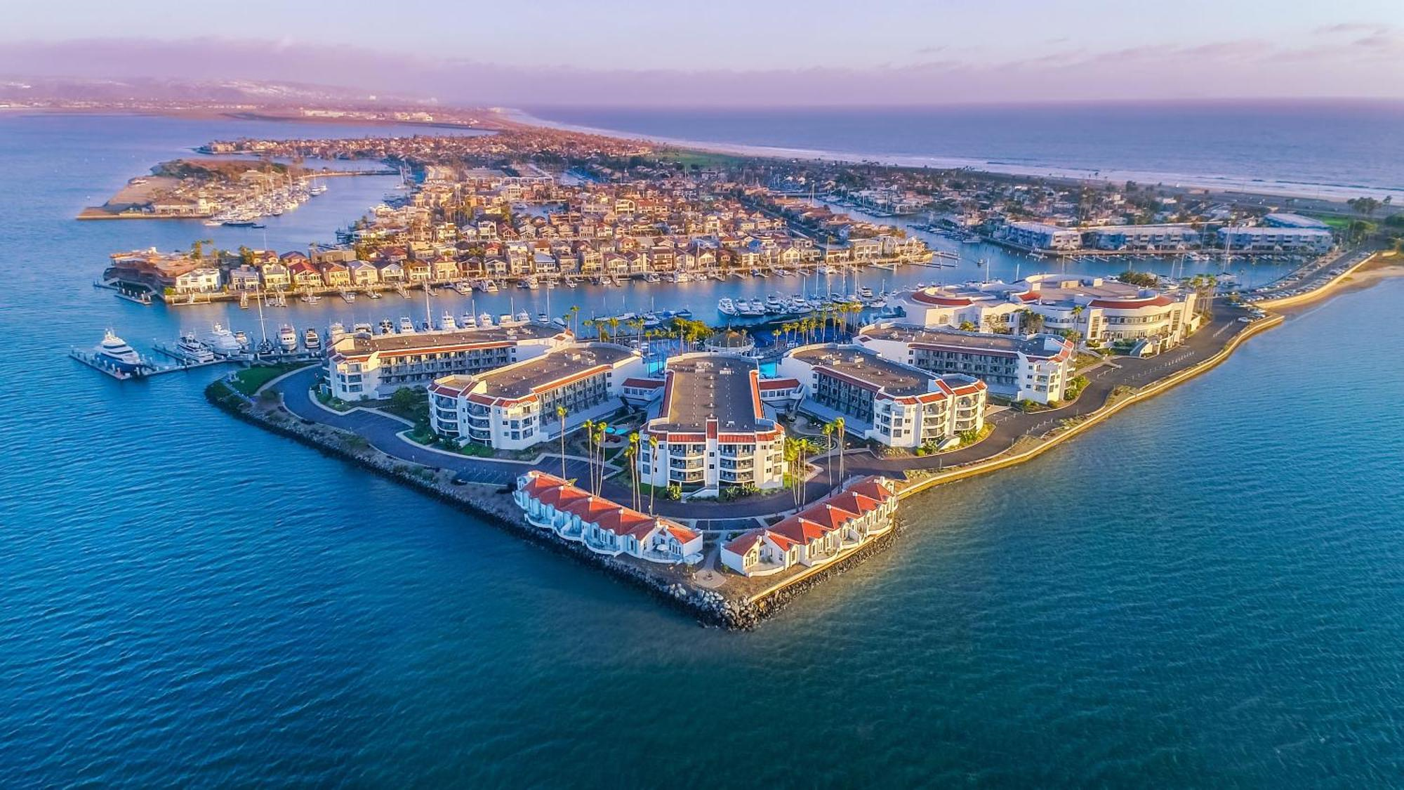 Aerial view of waterfront buildings and houses on a peninsula surrounded by water, with a marina and coastline visible in the background.
