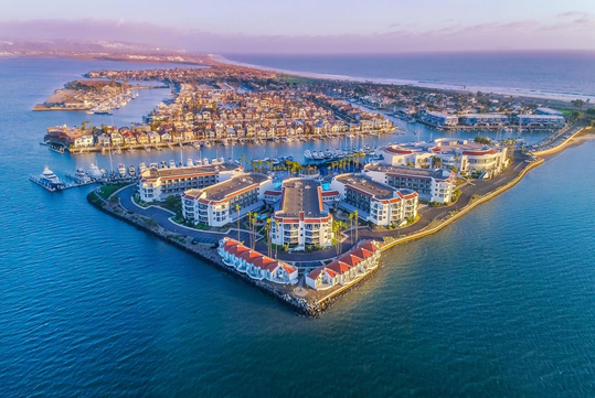 Aerial view of waterfront buildings and houses on a peninsula surrounded by water, with a marina and coastline visible in the background.
