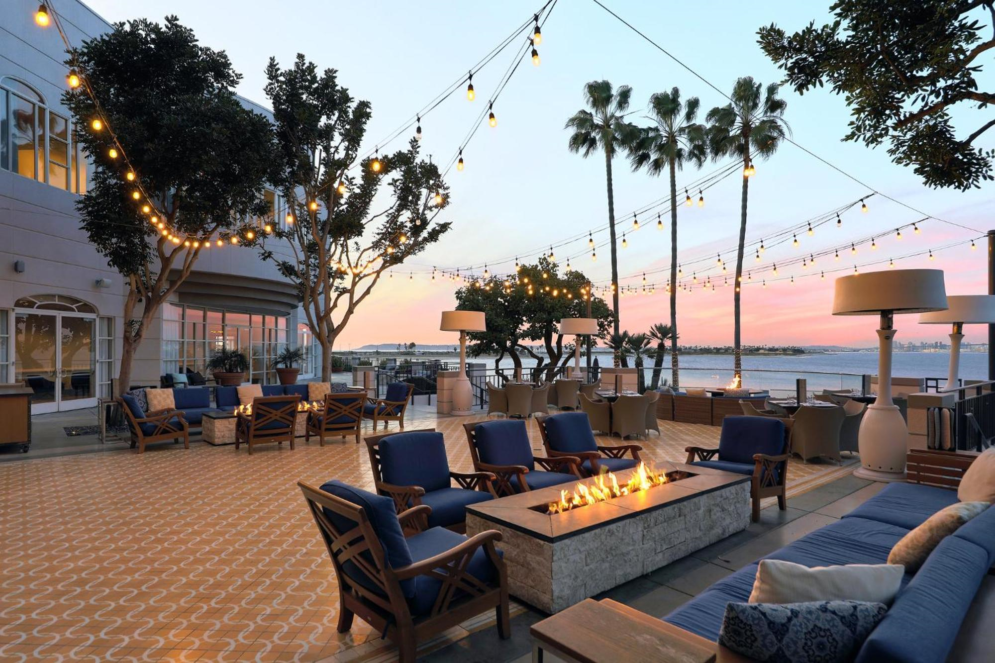 Outdoor patio with blue cushioned seating, a rectangular fire pit, string lights overhead, palm trees, and a waterfront view at sunset.