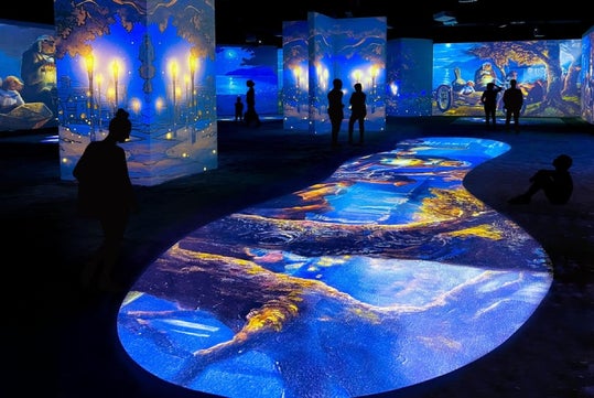 People view immersive digital art projections in a dark exhibition space featuring vibrant blue and yellow landscapes.