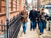 Four adults walk down a city sidewalk next to a brick building. One person carries a camera, and all are wearing coats. The street is lined with buildings and parked cars.