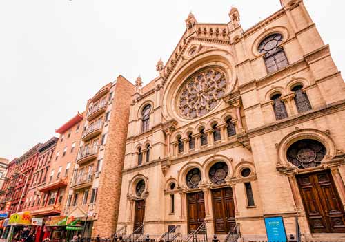 A historic stone synagogue with arched doorways and elaborate rose window, located on a city street with adjacent apartment buildings and shops.