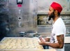 A man wearing a red hat and white shirt shapes dough on a tray in a commercial kitchen.
