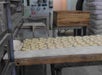 Rows of unbaked bagels are arranged on a floured work surface in a bakery, with baking trays and flour sacks in the background.