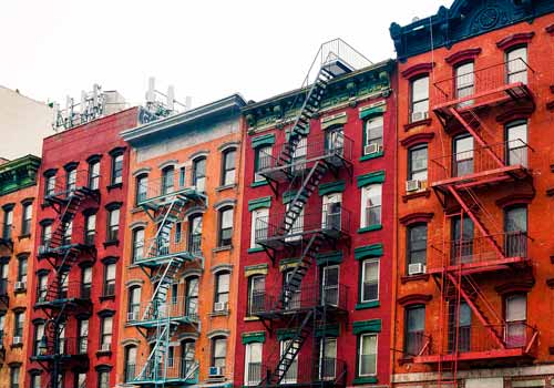 A row of colorful brick apartment buildings with fire escapes on their facades in an urban setting.
