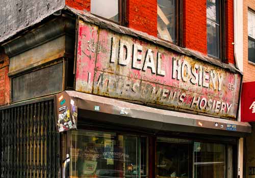 A weathered storefront sign reads "Ideal Hosiery" with faded lettering above a glass window and a metal security gate on an urban street.