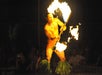 A performer in a grass skirt spins a flaming torch, creating trails of fire during a fire dance in front of an audience.