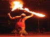 A performer with tattoos and grass leg bands balances a flaming stick on his mouth during a fire dancing act.