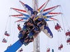 People are riding an amusement park plane-themed ride, with a second swing ride visible in the background against a clear sky.