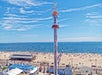 A tall amusement park ride towers over a crowded sandy beach with people sunbathing and swimming, set against a blue sky and ocean.