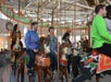 Several adults ride colorful carousel horses indoors, smiling and talking as the carousel spins. The background shows lights, poles, and parts of other horses.