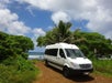 A white passenger van parked on a dirt road on a sunny day with trees and the ocean behind it.