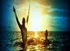 Two people are in the ocean at sunset, one with arms raised and water splashing, the other standing nearby, both silhouetted against the bright sky.