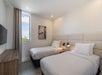 Two neatly made twin beds in a modern room with light-colored walls, a patterned accent wall, a wall-mounted TV, a window with curtains, and a small bedside lamp.