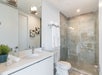 Modern bathroom with glass shower, white vanity, toilet, and gray tile. Towels hang near shower. Two decorative artworks and a potted plant adorn the space.