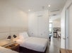 A modern bedroom with two single beds, bedside tables with lamps, patterned wallpaper, and a view into a kitchen and dining area.
