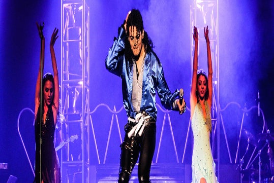 A Michael Jackson impersonator in a shiny blue shirt with back up dancers on either side of him on stage.