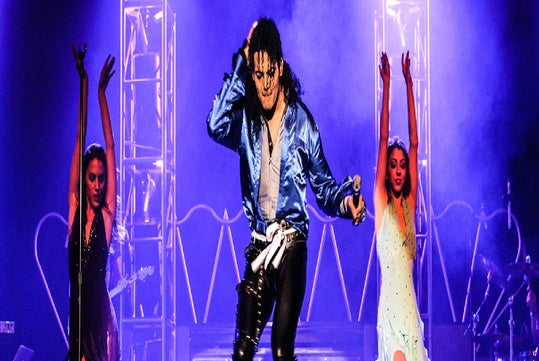 Performer in a blue jacket and black pants holding a microphone on stage, flanked by two dancers in black and white dresses against a purple-lit backdrop.