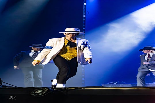 Performer in a white hat and jacket dances on stage with others in suits under blue lighting.