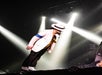 Performer in white suit and hat leans forward on stage, illuminated by spotlights.