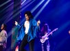 Performer in a blue jacket holds a microphone, dancing on stage with a guitarist and another person. Blue and purple stage lights illuminate the scene.