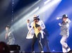 Performers on stage in suits and hats, with one in a white jacket and beige hat dancing at the center, surrounded by blue and purple lighting.