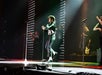 Performer dancing energetically on stage with backup dancers and a musical band in the background, under bright stage lights.