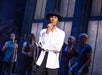 A person in a white shirt and black hat stands pensively on stage, with performers blurred in the background.