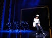 Performers on stage in a dramatic pose, with one in the foreground wearing a white hat and black-and-white outfit, and several others in the background wearing black outfits under blue lighting.