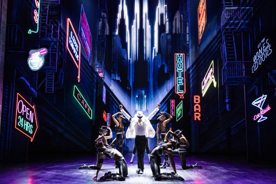 Performer in white hat and clothes dances center stage, surrounded by dancers in black outfits. Neon signs for hotels, bars, and cafes illuminate the urban alley backdrop.