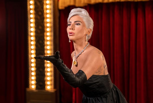 A wax figure of Lady Gaga, dressed in an elegant black gown, standing on a stage with a rich red curtain and ornate lighting at Madame Tussauds Hollywood.