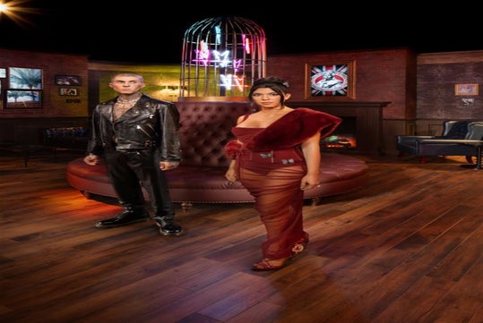A man in a leather outfit and a woman in a red dress stand in a dimly lit room with a round sofa and neon lights.