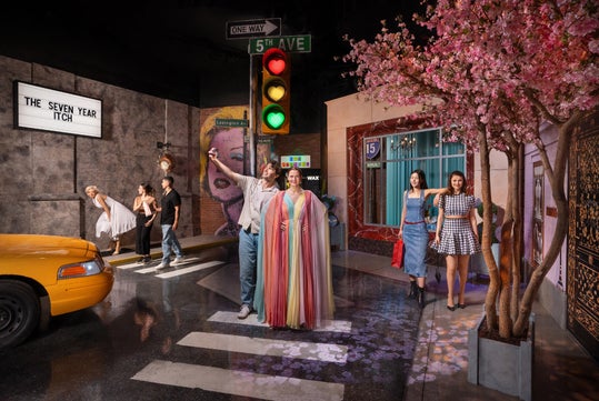 People pose in a street scene set, featuring iconic movie references, a yellow taxi, a traffic light, and a pink blossom tree, under a sign reading "The Seven Year Itch.